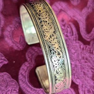 Könnte beinhalten: Silbernes Armreif-Armband mit aufwendigem, durchbrochenem Blumen- und Scroll-Design. Das Armband hat ein detailliertes Muster mit dunklem Hintergrund und geflochtenem Rand. Der Schmuck wird auf einer strukturierten, lila Oberfläche präsentiert.