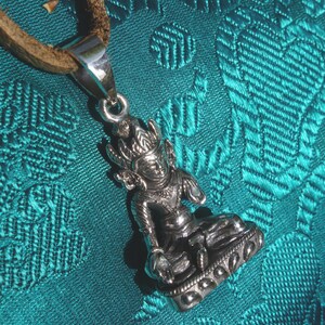 May include: Silver pendant necklace featuring a detailed sculpture of a seated Buddha figure with intricate details. The pendant is on a brown leather cord.