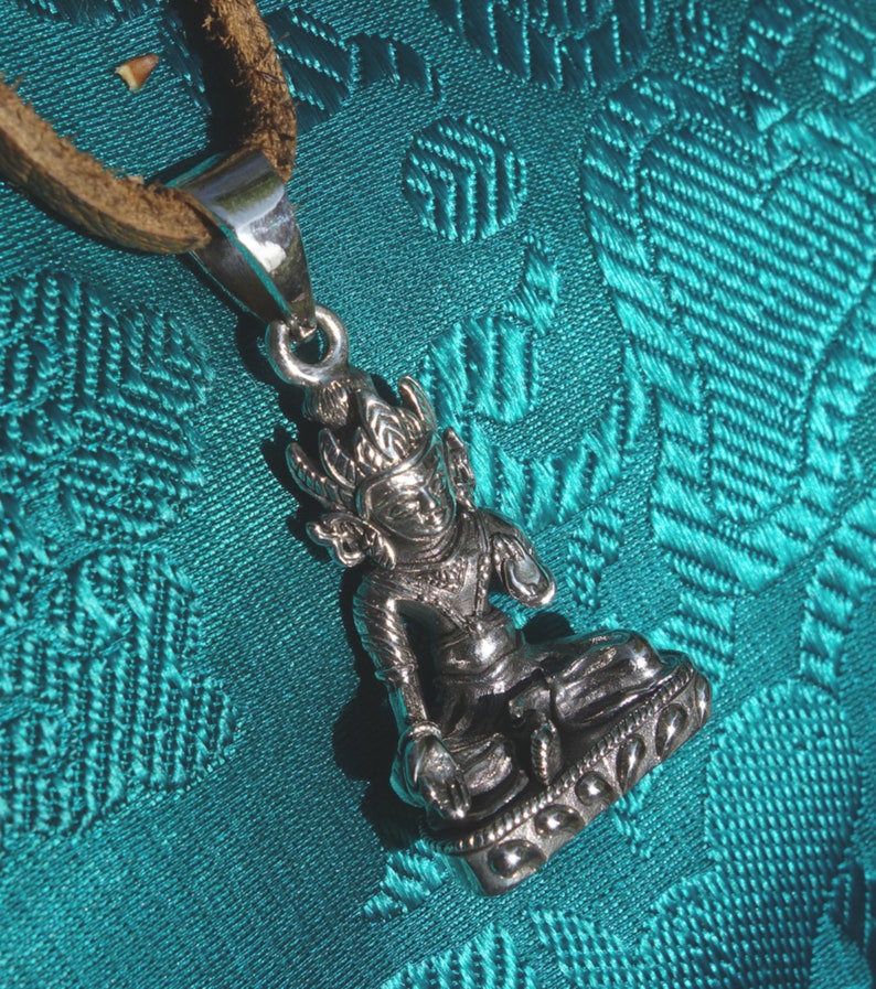 May include: Silver pendant necklace featuring a detailed sculpture of a seated Buddha figure with intricate details. The pendant is on a brown leather cord.