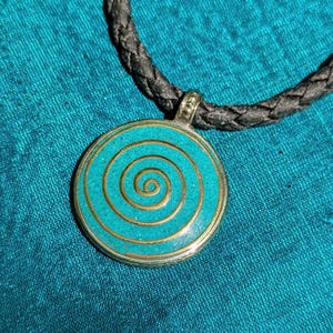 Op de afbeelding: Een ronde hangerketting met een turquoise spiraalontwerp. De hanger heeft een zilverkleurige rand en een goudkleurige spiraal. De ketting heeft een zwart gevlochten koord.