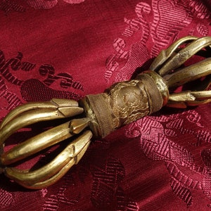 Op de afbeelding: Een gouden metalen handgehouden gebedswiel met een bruine leren wikkeling. Het gebedswiel heeft een decoratief ontwerp en bevindt zich op een rode stoffen achtergrond.