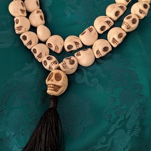 May include: A necklace with skull-shaped beads in off-white, arranged in multiple strands. It has a black tassel and is set against teal patterned fabric. The skulls are intricately carved, creating a unique design.