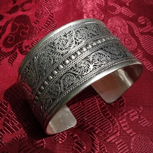 Op de afbeelding: Zilveren manchetarmband met ingewikkelde filigraan- en kralenaccenten. De brede band heeft bloemmotieven en een gepolijste afwerking. Het open ontwerp maakt het gemakkelijk om te dragen. De armband wordt getoond op een rood patroonweefsel.