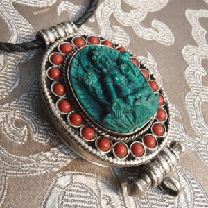 Op de afbeelding: Een zilveren ketting met een hanger van groen gegraveerd materiaal en een rand van rode koraal kralen. De ketting ligt op een bruine stoffen achtergrond.