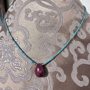Puede incluir: Un collar turquesa y plateado con un colgante de piedra roja en forma de lágrima.
