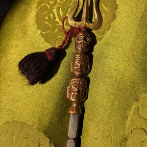 May include: A brass trident pendant with a skull and two carved faces, attached to a silver and grey beaded chain. A dark tassel with red accents hangs from the pendant. The pendant is displayed against a green background.