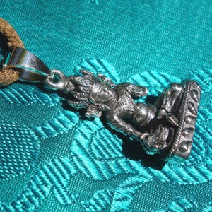 May include: Silver pendant necklace featuring a detailed depiction of a seated Buddha figure with a crown and intricate details. The pendant is attached to a brown leather cord.