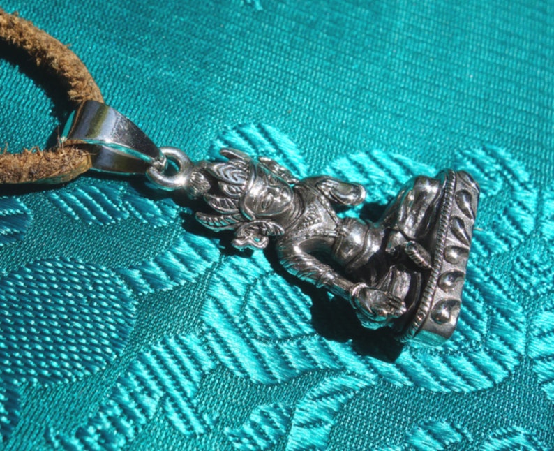 May include: Silver pendant necklace featuring a detailed depiction of a seated Buddha figure with a crown and intricate details. The pendant is attached to a brown leather cord.