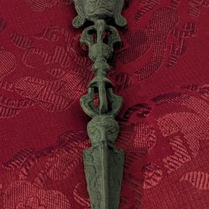 May include: A dark green metal dagger with a decorative handle featuring a face and intricate designs. The dagger is on a red fabric background.