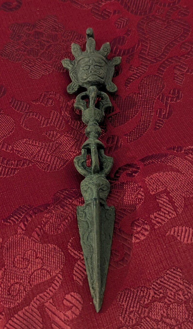 May include: A dark green metal dagger with a decorative handle featuring a face and intricate designs. The dagger is on a red fabric background.