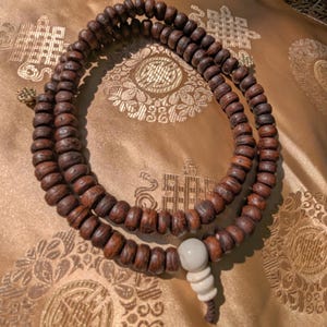 Peut inclure: Un chapelet en bois brun foncé, également appelé mala, avec un pendentif blanc et crème. Les perles sont disposées en cercle. Le fond est un tissu doré avec un motif répétitif.