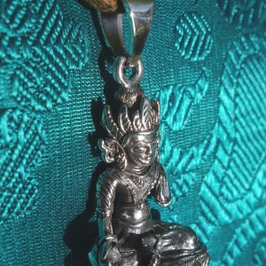 May include: A silver pendant featuring a detailed depiction of a seated figure with a crown and ornate clothing. The pendant is attached to a brown leather cord.