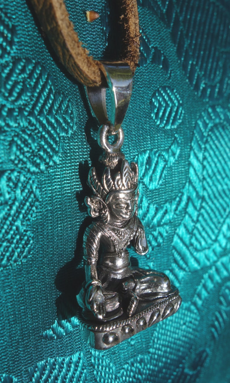 May include: A silver pendant featuring a detailed depiction of a seated figure with a crown and ornate clothing. The pendant is attached to a brown leather cord.