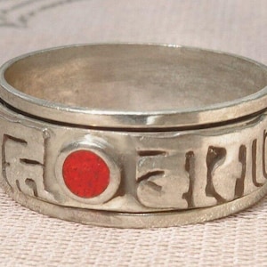 Könnte beinhalten: Ein drehbarer Silberring mit einem roten Stein und gravierter Beschriftung.