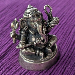 May include: A silver-coloured statue of Ganesha, the Hindu god of wisdom and prosperity, with four arms and a trunk. The statue is sitting on a round base and is decorated with intricate details.