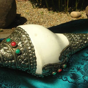May include: A white conch shell with ornate silver metalwork. The metalwork is detailed with red and green gemstones. The shell rests on a teal fabric with a dragon pattern. The conch shell is a decorative item.