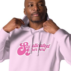Może przedstawiać: Jasnoróżowa bluza z kapturem z przednią kieszenią i kapturem ze sznurkiem. Bluza z kapturem ma napis "Sophisticated Expert" w stylu retro różową czcionką. Osoba ma na sobie bluzę z kapturem.