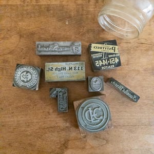 May include: A collection of vintage printing stamps, including rectangular and circular designs, with text and logos. The stamps are made of metal and wood, and are arranged on a wooden surface. A clear glass jar is in the background.