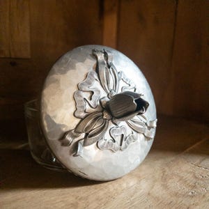 May include: A silver-colored jar lid with a hammered texture and a floral design. The lid features a raised flower and ribbon motif. The jar is partially visible, made of clear glass, and sits on a wooden surface.
