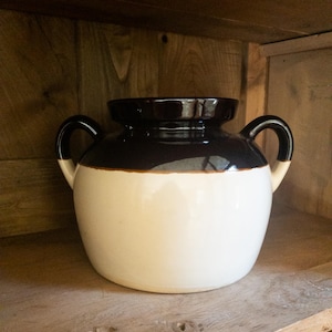 May include: A two-toned ceramic jar with a dark brown top and handles, and a cream-colored base. The jar has two curved handles and sits on a wooden shelf. The jar is a decorative item.