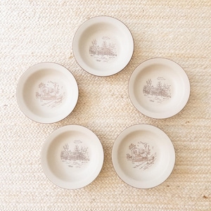 May include: Five off-white ceramic plates with a brown rim and a detailed brown landscape scene in the center. The plates are arranged in a circular pattern, showcasing a vintage aesthetic. Each plate features a unique illustration.