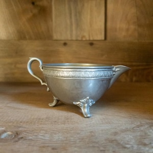 May include: A small, antique silver-colored gravy boat with a handle and spout. The boat rests on three ornate feet. The rim features a decorative pattern. The gravy boat is displayed on a wooden surface.