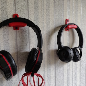 May include: Two pairs of black headphones with red accents hanging on red plastic wall mounts. The headphones are on a white textured wall.