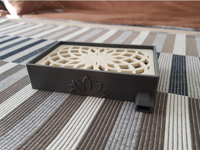 Puede incluir: Un quemador de incienso rectangular gris con un dise&ntilde;o de rejilla blanca decorativa en la parte superior. El quemador tiene un dise&ntilde;o de flor de loto en la parte delantera y un peque&ntilde;o agujero en el lateral.