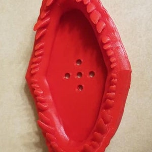 May include: Red plastic 3D printed dinosaur mouth shaped dish with a row of teeth around the edge. The dish has a series of small holes in the bottom.