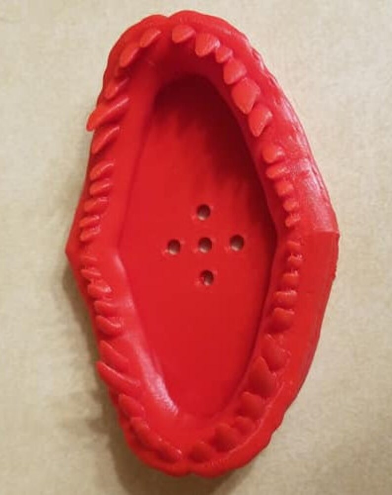 May include: Red plastic 3D printed dinosaur mouth shaped dish with a row of teeth around the edge. The dish has a series of small holes in the bottom.