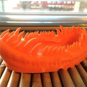 May include: Orange 3D printed bowl with a jagged, spiky rim. The bowl is sitting on a woven bamboo surface.