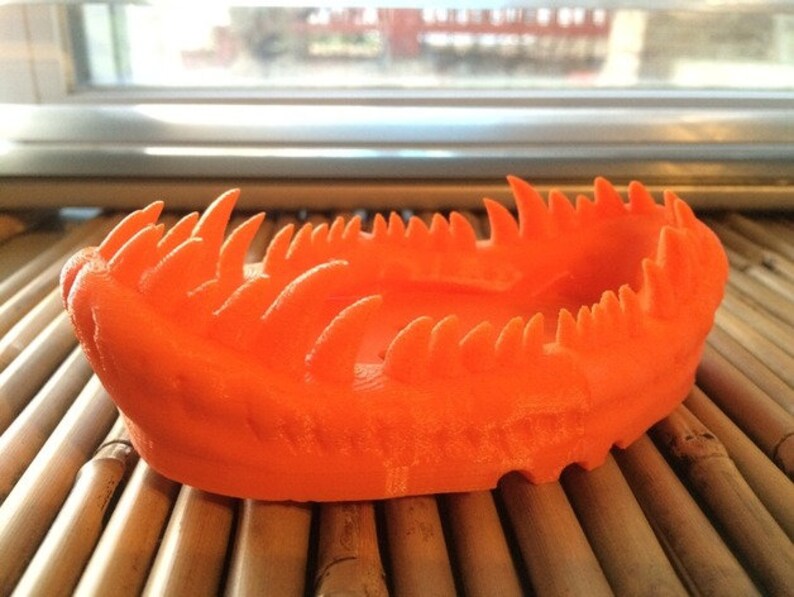 May include: Orange 3D printed bowl with a jagged, spiky rim. The bowl is sitting on a woven bamboo surface.
