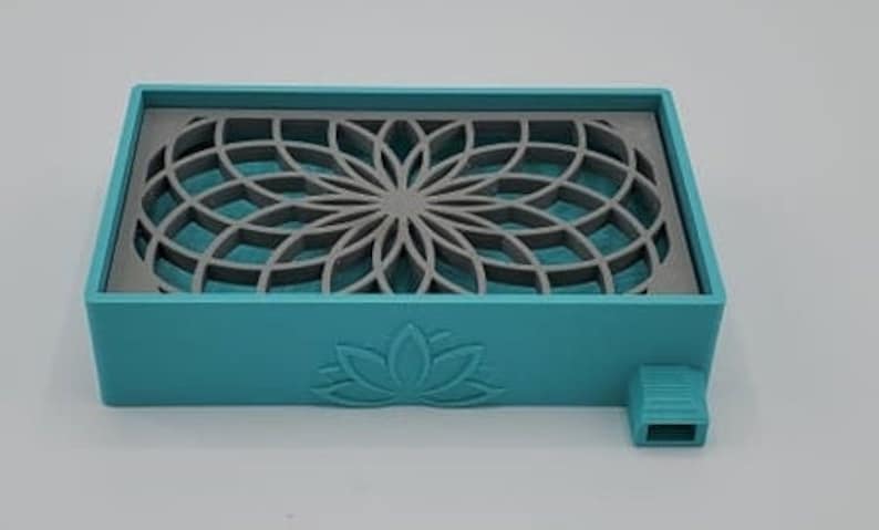Puede incluir: Un jabonera rectangular de color azul turquesa con un dise&ntilde;o geom&eacute;trico gris y un logotipo de flor de loto. La jabonera tiene una peque&ntilde;a abertura en el lateral.