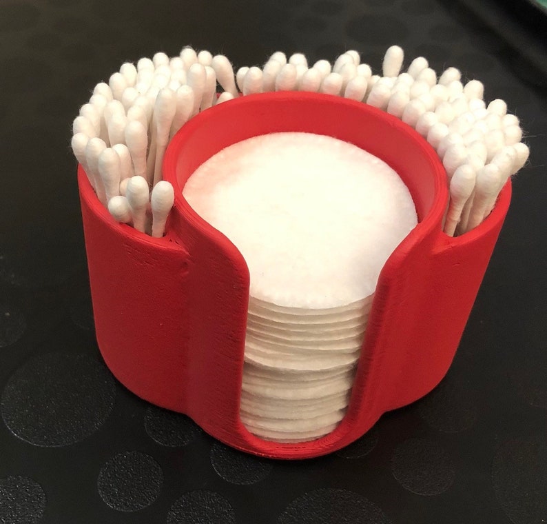 May include: Red plastic organizer with two compartments for storing cotton swabs and cotton rounds. The organizer has a rounded shape and is filled with white cotton swabs and cotton rounds.
