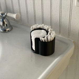 May include: A black plastic bathroom organizer with two compartments. One compartment holds cotton rounds and the other holds cotton swabs.