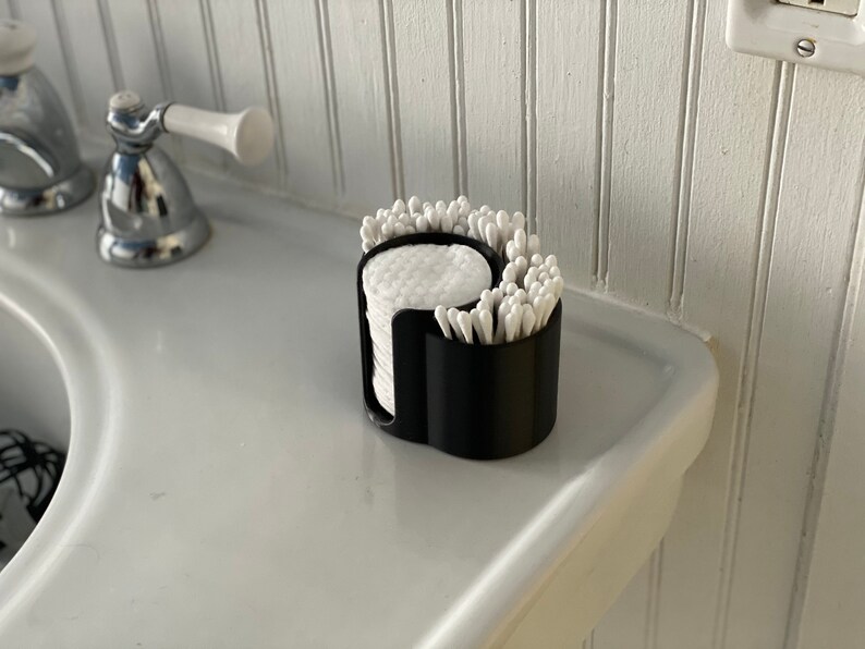 May include: A black plastic bathroom organizer with two compartments. One compartment holds cotton rounds and the other holds cotton swabs.