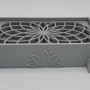 Puede incluir: Una caja gris impresa en 3D con un dise&ntilde;o geom&eacute;trico en la parte superior y un dise&ntilde;o de flor de loto en la parte delantera. La caja tiene un peque&ntilde;o pico en el lateral.