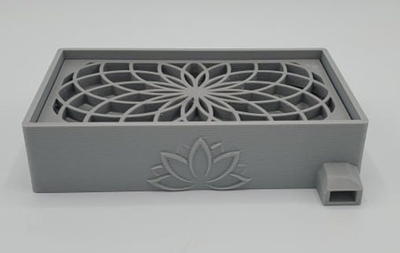 Puede incluir: Una caja gris impresa en 3D con un dise&ntilde;o geom&eacute;trico en la parte superior y un dise&ntilde;o de flor de loto en la parte delantera. La caja tiene un peque&ntilde;o pico en el lateral.