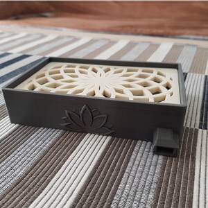 Puede incluir: Un quemador de incienso rectangular gris con un dise&ntilde;o de rejilla blanca decorativa en la parte superior. El quemador tiene un dise&ntilde;o de flor de loto en la parte delantera y un peque&ntilde;o agujero en el lateral.