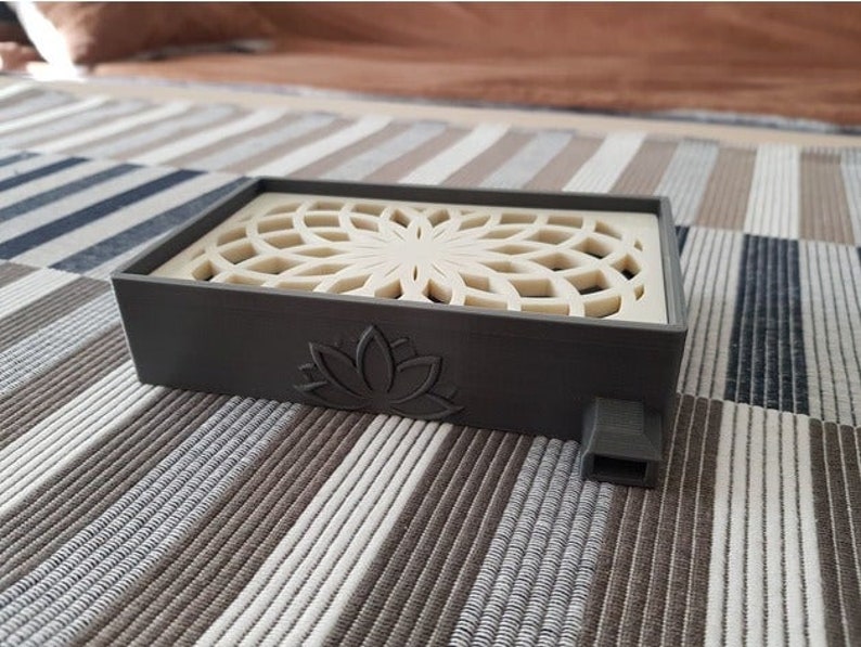 Puede incluir: Un quemador de incienso rectangular gris con un dise&ntilde;o de rejilla blanca decorativa en la parte superior. El quemador tiene un dise&ntilde;o de flor de loto en la parte delantera y un peque&ntilde;o agujero en el lateral.