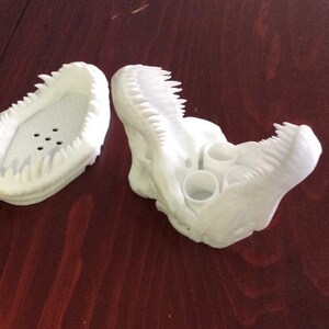 May include: A white 3D printed dinosaur head with an open mouth and a row of teeth. The dinosaur head has three holes in the top for holding pens or pencils.