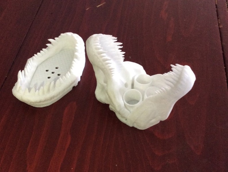 May include: A white 3D printed dinosaur head with an open mouth and a row of teeth. The dinosaur head has three holes in the top for holding pens or pencils.