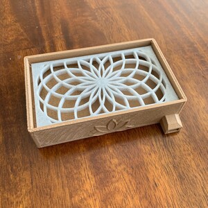 Puede incluir: Una caja de madera marr&oacute;n claro con un dise&ntilde;o geom&eacute;trico blanco en la parte superior. La caja tiene una peque&ntilde;a abertura en el lateral y un dise&ntilde;o de flor de loto en la parte inferior.