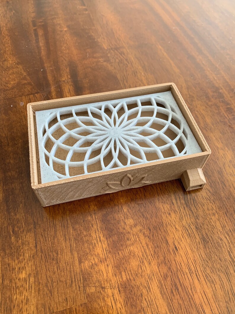 Puede incluir: Una caja de madera marr&oacute;n claro con un dise&ntilde;o geom&eacute;trico blanco en la parte superior. La caja tiene una peque&ntilde;a abertura en el lateral y un dise&ntilde;o de flor de loto en la parte inferior.