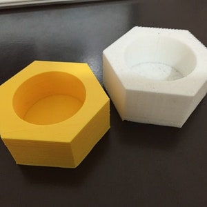 May include: Two hexagonal, open-topped containers. One is bright yellow, the other is white. Both have a circular opening in the center. The containers are likely 3D-printed and sit on a dark surface.