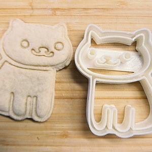 Puede incluir: Dos cortadores de galletas de plástico blanco con forma de gato. Los cortadores están sobre una superficie de madera.