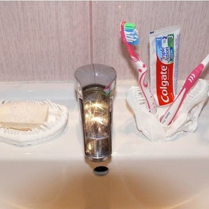 May include: A white ceramic dinosaur skull toothbrush holder with two pink toothbrushes and a tube of Colgate Triple Action toothpaste.