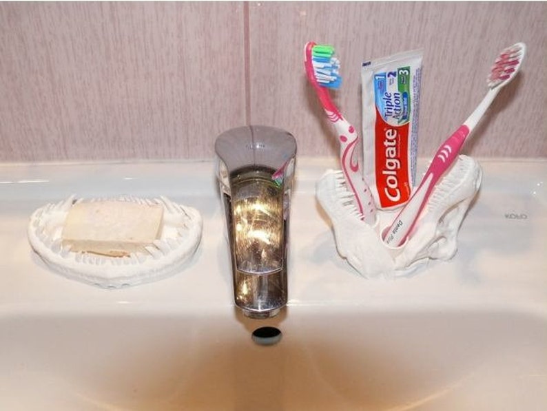 May include: A white ceramic dinosaur skull toothbrush holder with two pink toothbrushes and a tube of Colgate Triple Action toothpaste.