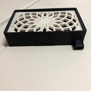 Puede incluir: Una caja rectangular negra con un dise&ntilde;o geom&eacute;trico blanco en la parte superior y un dise&ntilde;o de flor de loto en la parte delantera. La caja tiene una peque&ntilde;a boquilla negra en el lateral.