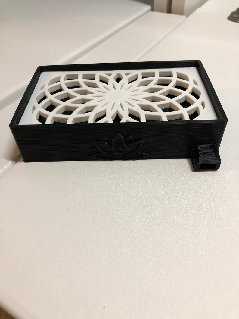 Puede incluir: Una caja rectangular negra con un dise&ntilde;o geom&eacute;trico blanco en la parte superior y un dise&ntilde;o de flor de loto en la parte delantera. La caja tiene una peque&ntilde;a boquilla negra en el lateral.